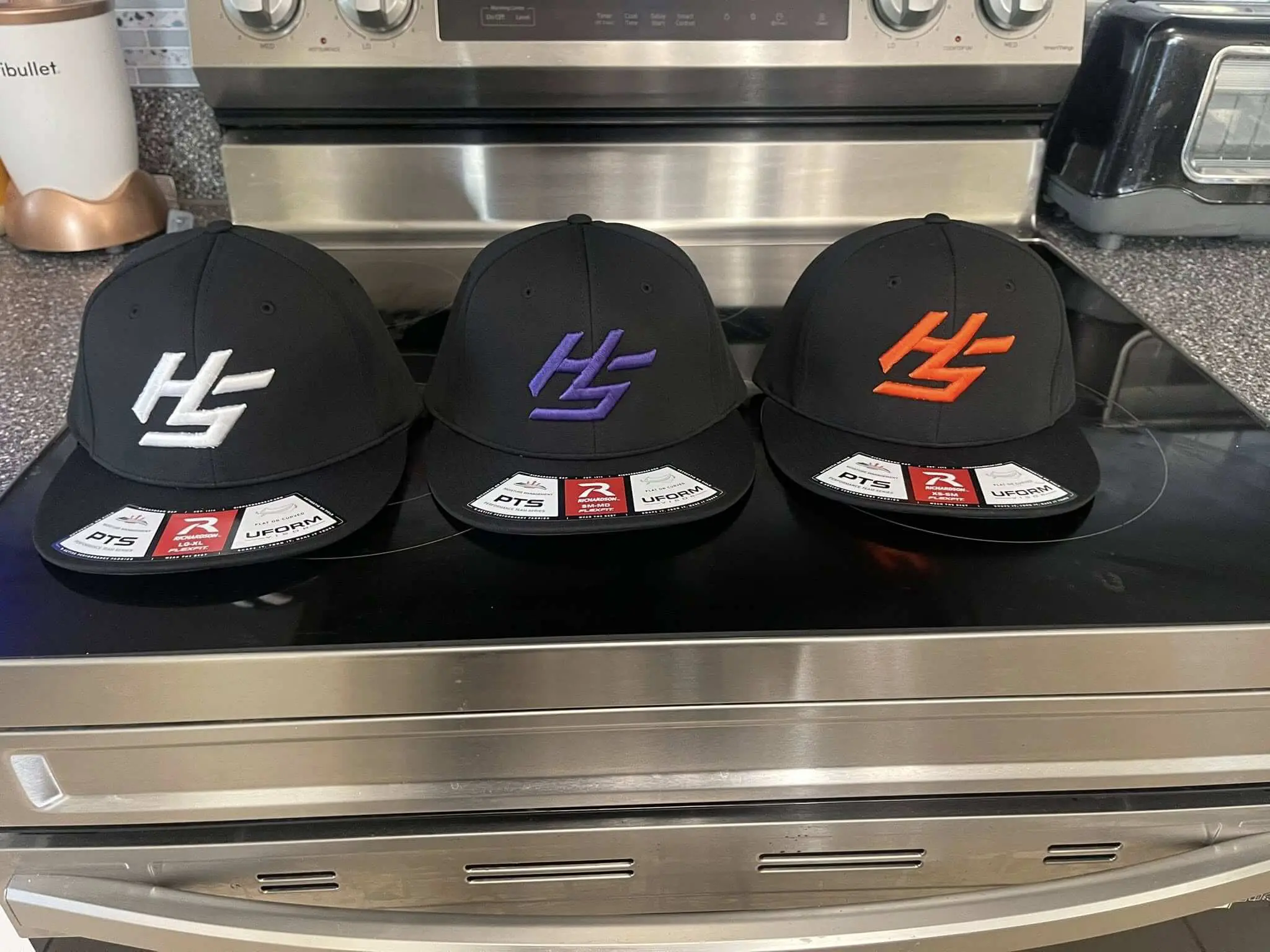 Three black baseball caps with different colored logos on a kitchen counter.