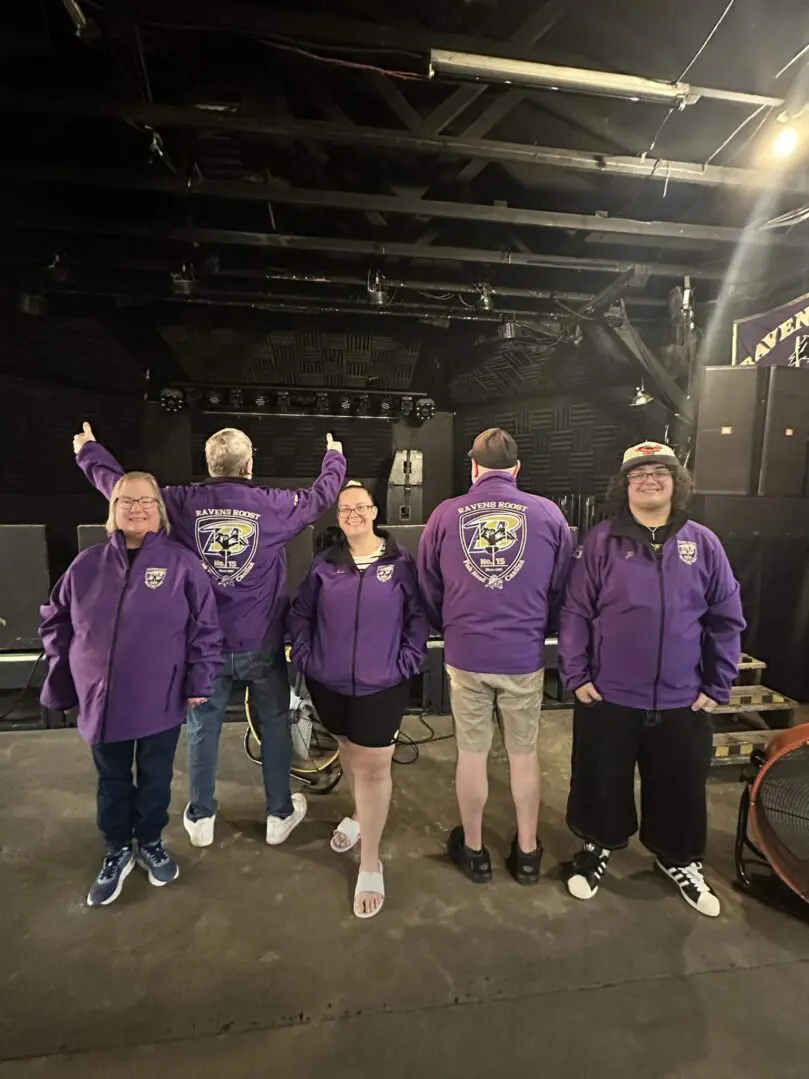 Group of five people wearing purple shirts and party hats indoors.