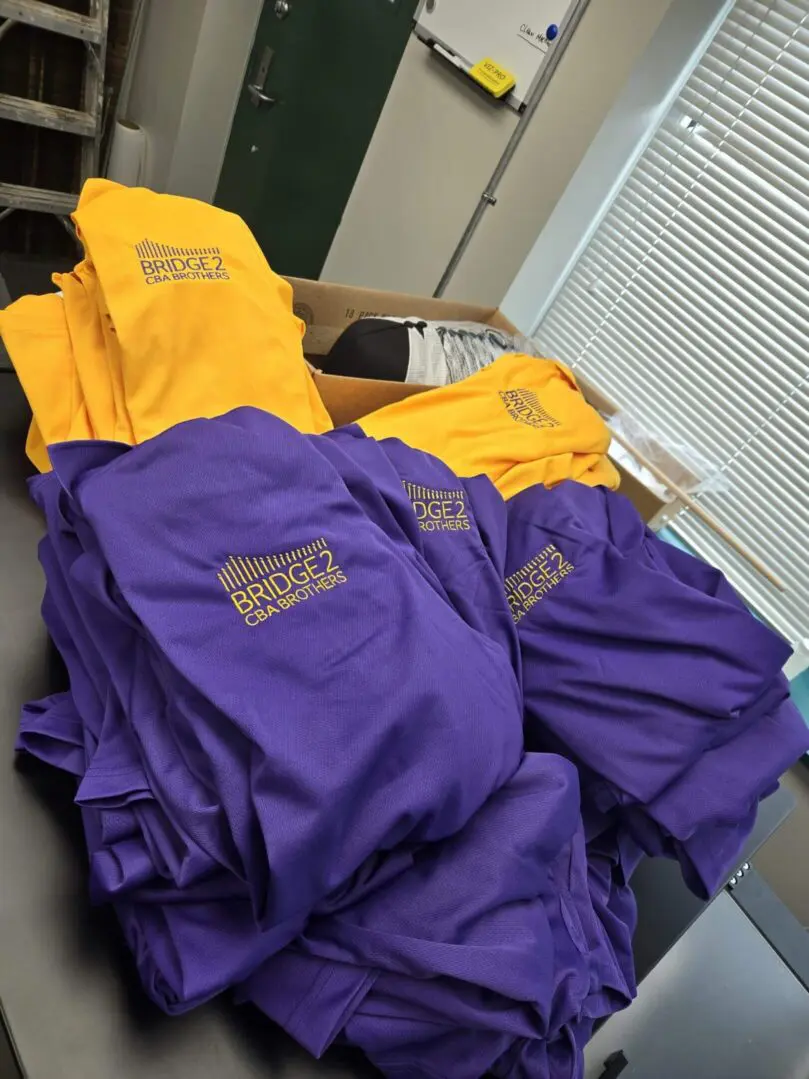 Stacks of purple and yellow uniforms with embroidered logos on a table.